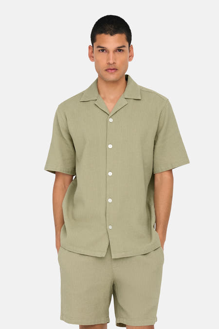 Chemise beige à manches courtes de Only & Sons, avec col boutonné, portée avec des shorts coordonnés, des chaussettes rayées et des baskets blanches.