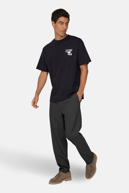 Man met zwart t-shirt met logo, grijze pantalon en bruine schoenen, staand.