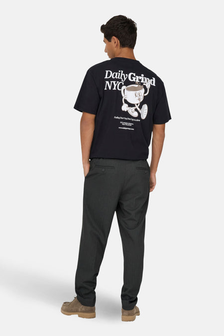Man met zwart T-shirt met "Daily Grind NYC" en cartoon, grijze broek en bruine schoenen, achterkant.