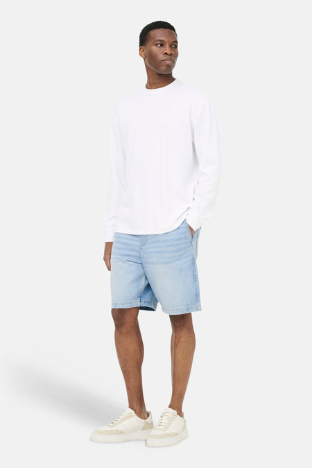Lichtblauwe denim jeansshort van ONLY & SONS®, gecombineerd met een wit shirt met lange mouwen en crèmekleurige sneakers.
