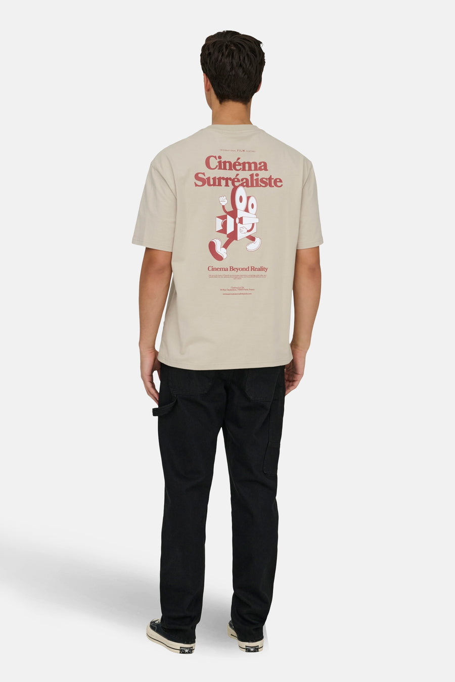T-shirt écru à manches courtes de ONLY & SONS®, avec une impression 'Cinéma Surréaliste' sur le dos, porté avec un pantalon noir et des baskets.