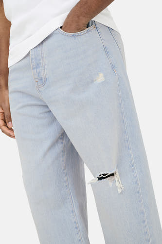 Jean large bleu clair en denim ONLY & SONS® avec déchirures, une main dans la poche et un t-shirt blanc.