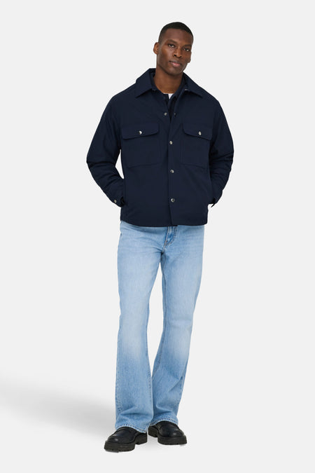 Man met marineblauw jasje en lichtblauwe jeans, staand.