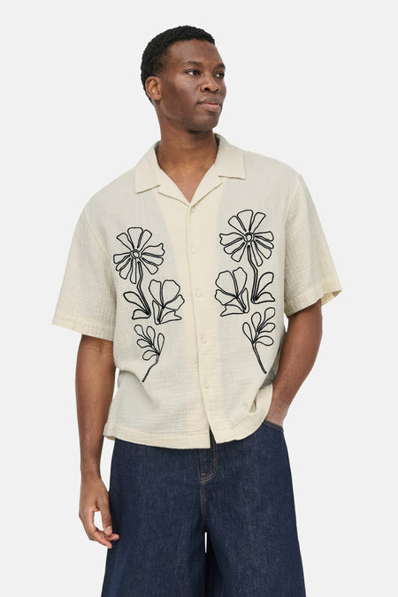 Chemise écru à manches courtes ONLY & SONS® avec imprimé floral noir, portée par un homme avec un short bleu foncé et des sandales beiges.