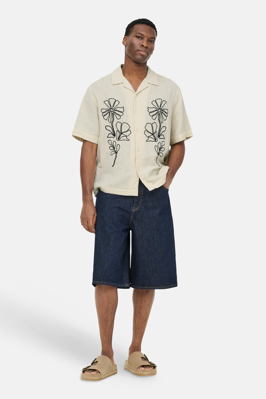 Ecru overhemd met korte mouwen van ONLY & SONS® met zwarte bloemenprint, gedragen door een man met een donkerblauwe korte broek en beige sandalen.