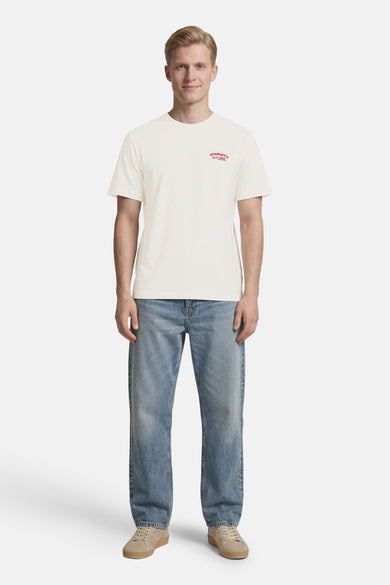 T-shirt de couleur crème de ONLY & SONS® avec petit imprimé rouge sur la poitrine, porté avec un jean bleu et des baskets beiges.