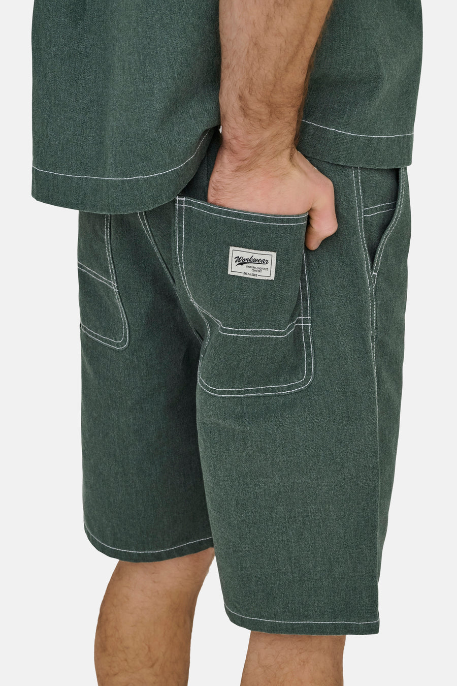 Short vert de ONLY & SONS® avec surpiqûres contrastées blanches, une texture et une étiquette sur la poche arrière, porté avec la main dans la poche.