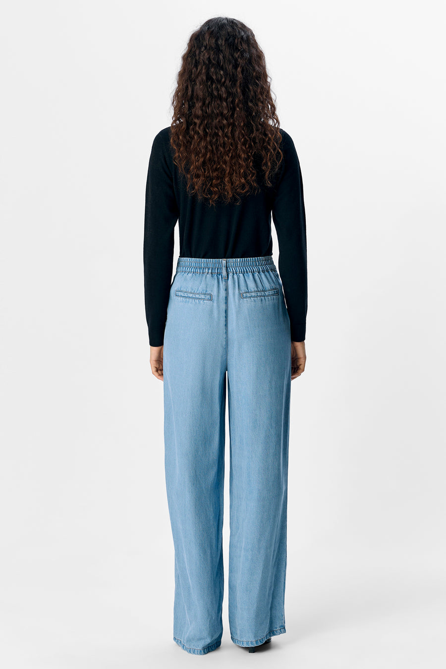 Achterkant van een vrouw met krullend haar, een zwarte trui en wijde, blauwe broek.