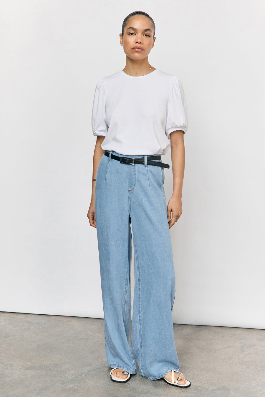 Vrouw in een witte top, wijde jeans met riem, en sandalen.