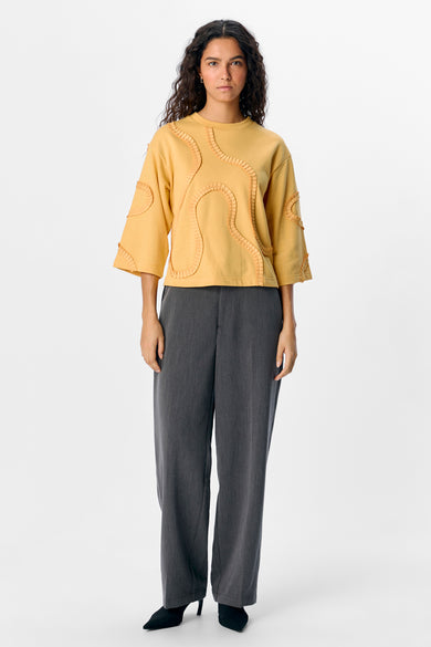 Pull-over jaune d'OBJET, avec des plis décoratifs, associé à un pantalon gris et des chaussures noires.