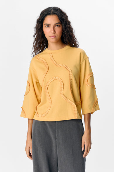 Pull-over jaune d'OBJECT, avec des applications décoratives et associé à un pantalon gris.