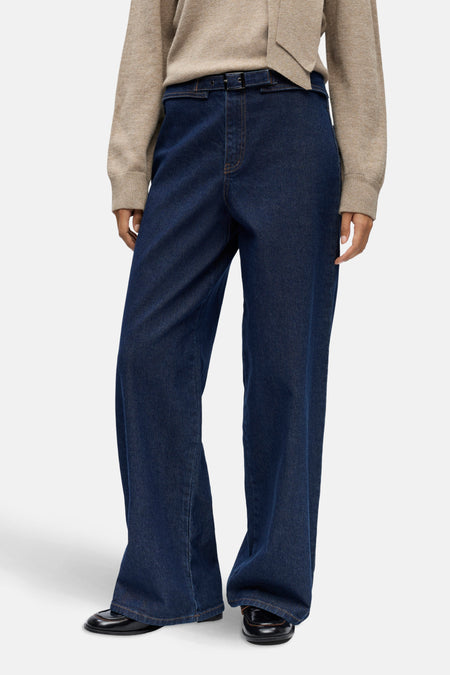 Model met beige trui en donkerblauwe wijde jeans met riem.