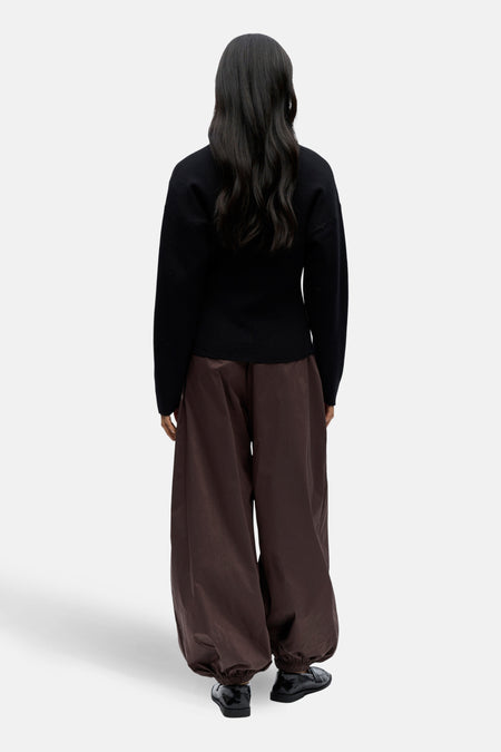 Achterkant van een vrouw in een zwarte trui en wijde, bruine broek, met zwarte schoenen op een witte achtergrond.
