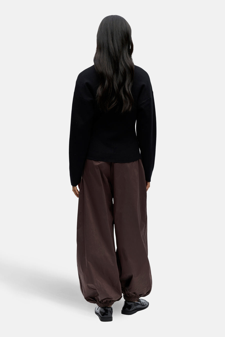 Achterkant van een vrouw in een zwarte trui en wijde, bruine broek, met zwarte schoenen op een witte achtergrond.
