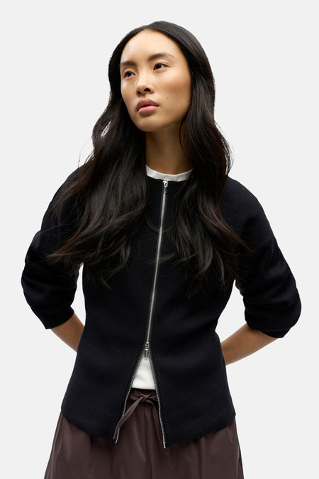 Vrouw met zwarte jas met rits, bruine broek, staand.