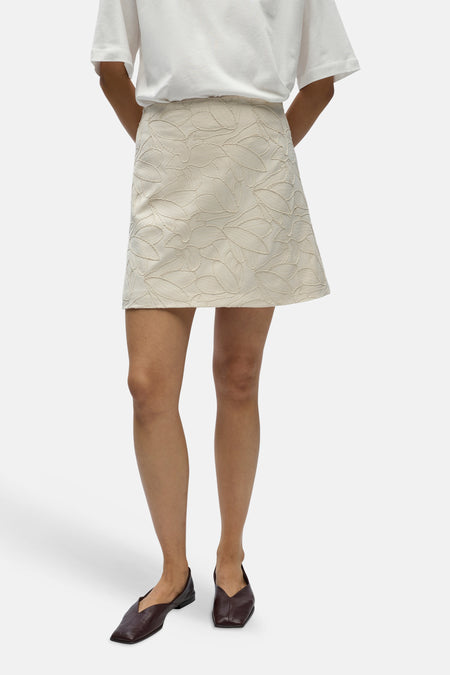 Jupe courte écru d'OBJECT, avec un motif de feuilles et combinée avec un t-shirt blanc et des chaussures plates marron.