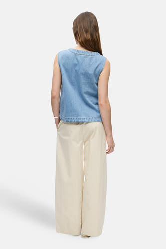 Chemisier sans manches en denim bleu clair de OBJECT, vue de dos du mannequin aux longs cheveux bruns, stylisé avec un pantalon large de couleur crème et un bracelet argenté.