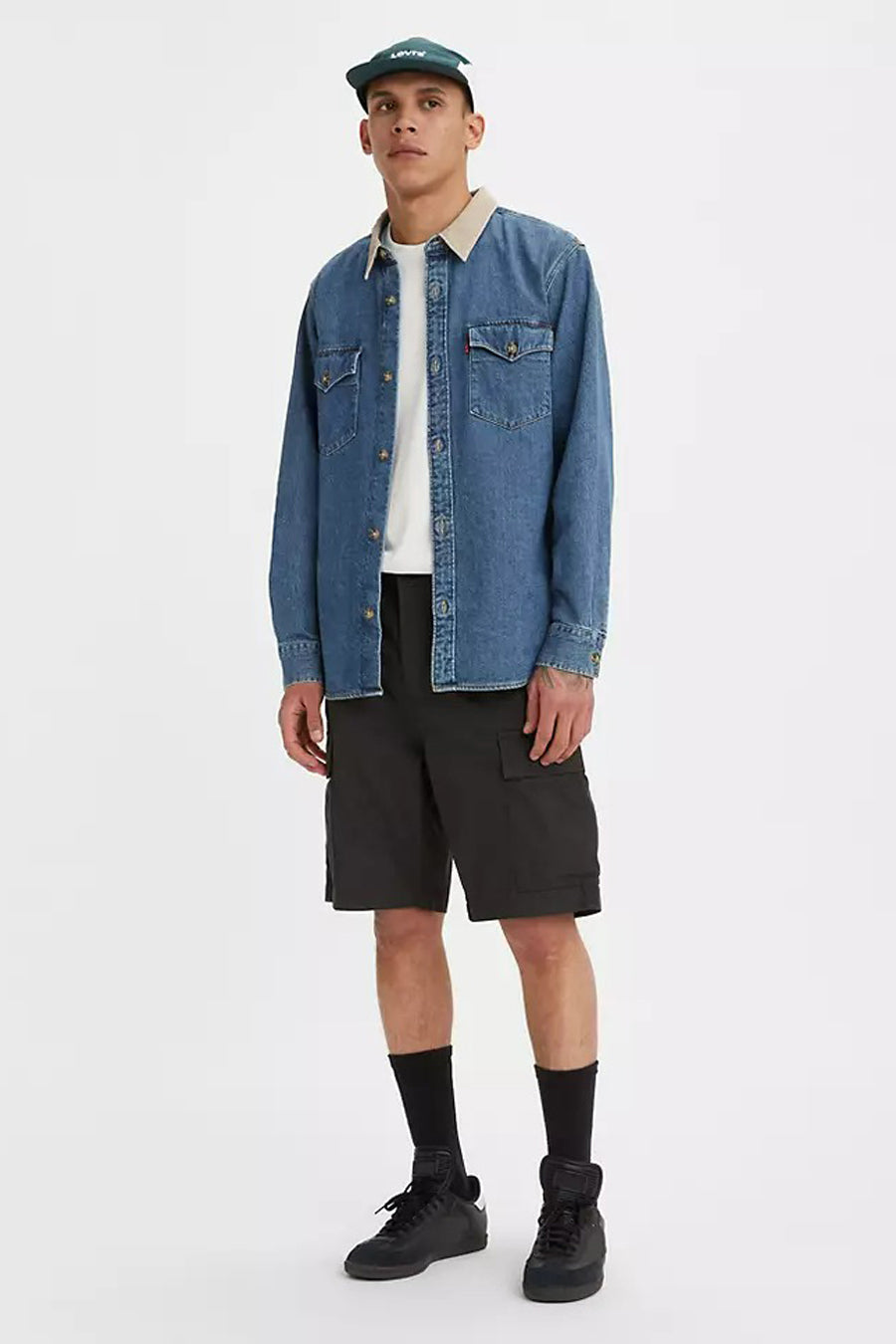 Man in jeanshemd, T-shirt, zwarte cargoshorts, sokken, sneakers en pet op witte achtergrond.