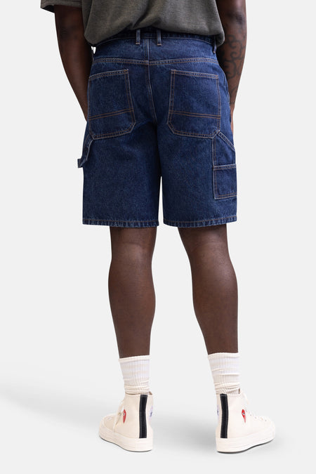 Short de denim bleu marine de REDEFINED REBEL, avec des poches arrière et une poche à outils, porté avec des chaussettes blanches et des baskets crème.