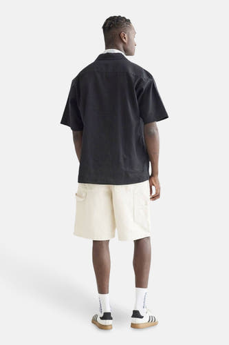 Chemise noire à manches courtes de WOODBIRD, vue de derrière, avec des shorts crème et des chaussures de sport.