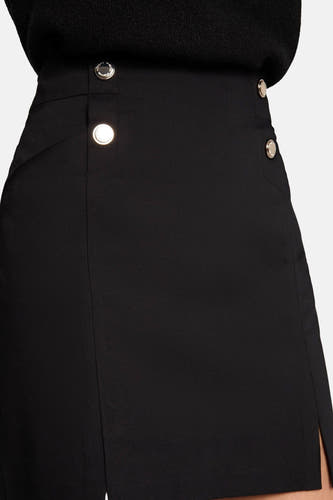 Jupe courte noire de Morgan De Toi, avec une fente et des boutons dorés. Portée avec un haut noir, une chaîne d'argent et des bottes noires.