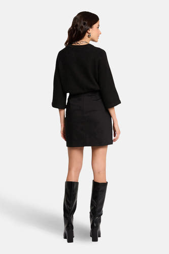 Jupe courte noire de Morgan De Toi, vue de derrière, associée à un pull noir et des bottes en cuir jusqu'aux genoux.