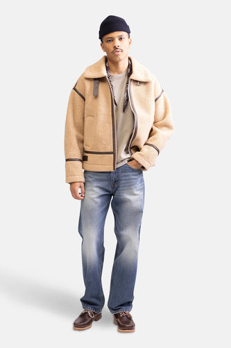 Man in jas met teddyvoering, jeans en boots tegen witte achtergrond.