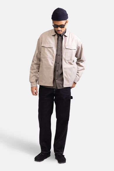 Chemise brune à longues manches de REDEFINED REBEL, portée sous un manteau beige avec un pantalon noir et un chapeau.
