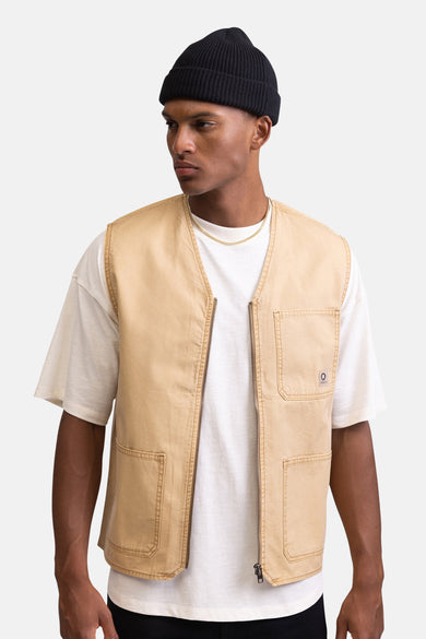 Manteau sans manches beige de REDEFINED REBEL, avec des boutons sur l'épaule, associé à un T-shirt blanc et une beanie noire.
