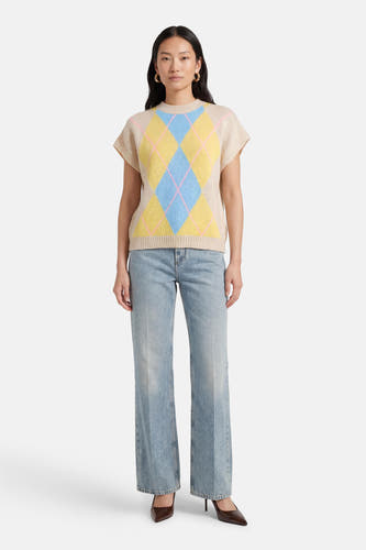 Beige mouwloos gilet van Terra di Siena, met een argyle-ruitpatroon in blauw en geel, gedragen over een jeans met wijde pijpen en bruine pumps.
