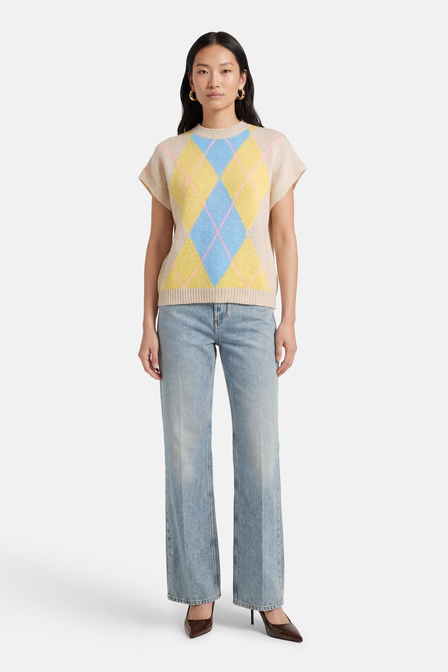 Beige mouwloos gilet van Terra di Siena, met een argyle-ruitpatroon in blauw en geel, gedragen over een jeans met wijde pijpen en bruine pumps.
