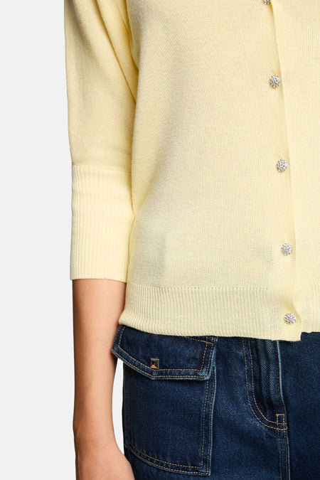 Cardigan Morgan De Toi en maille fine de couleur jaune avec manches trois-quarts, bords-côtes aux manches et à la base, et boutons scintillants, partiellement sur un jean bleu foncé.