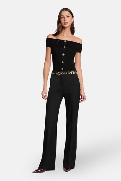 Pull-over noir de Morgan De Toi, avec les épaules découvertes et des boutons dorés, associé à une ceinture dorée et à un pantalon noir.