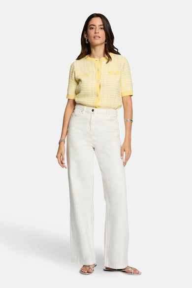 Cardigan jaune de Morgan de Toi, avec un motif de carreaux subtil et des boutons décoratifs, associé à une jupe blanche et des sandales argentées.