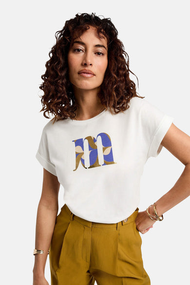 T-shirt blanc à manches courtes de Morgan De Toi, avec une grande impression bleue et dorée "M", associé à un pantalon marron et des bracelets d'or.