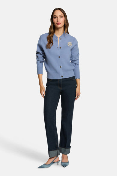 Gilet bleu de Morgan De Toi, avec fermeture à boutons et broche décorative, associé à des jeans foncés et des escarpins bleus.