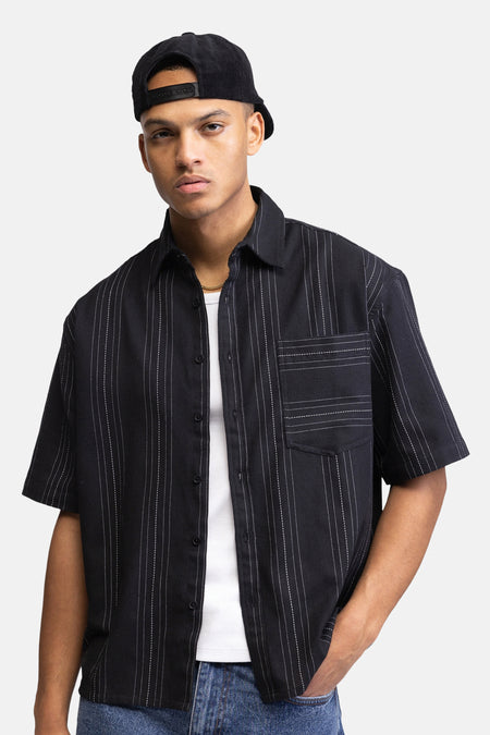 Chemise noire à manches courtes de REDEFINED REBEL, avec de fines rayures blanches, vue de dos sur un mannequin portant une casquette noire en velours côtelé et un jean bleu.