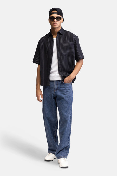 Chemise noire à manches courtes de REDEFINED REBEL, avec de fines lignes verticales blanches à pois, portée ouverte sur un t-shirt blanc et un jean bleu.