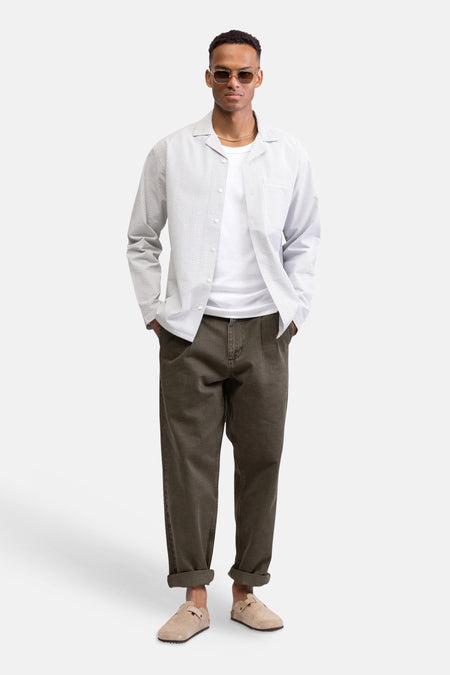 Chemise blanche à rayures à manches longues de REDEFINED REBEL, portée sur un t-shirt blanc, un pantalon marron et des sabots beiges.