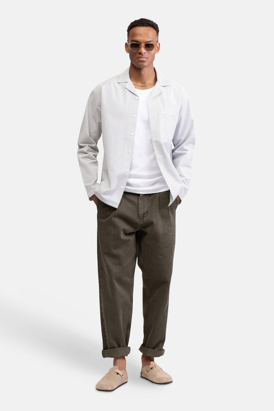 Chemise blanche à rayures à manches longues de REDEFINED REBEL, portée sur un t-shirt blanc, un pantalon marron et des sabots beiges.