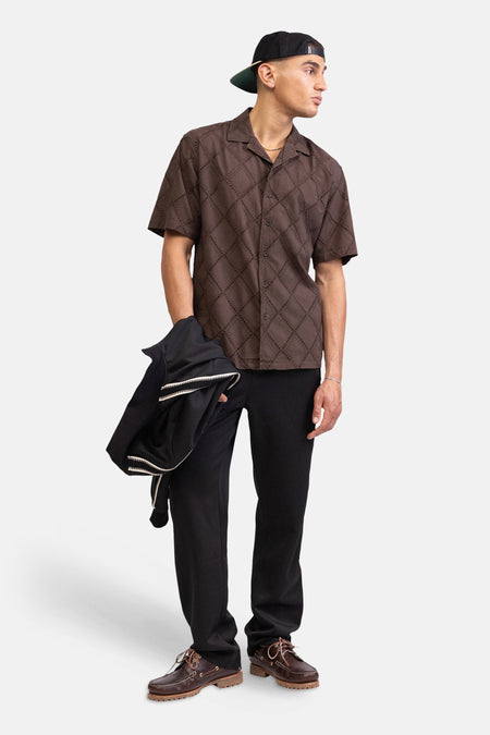 Chemise marron à manches courtes de REDEFINED REBEL, dotée d'un motif ajouré en losange, portée avec un pantalon noir et des chaussures bateau marron.