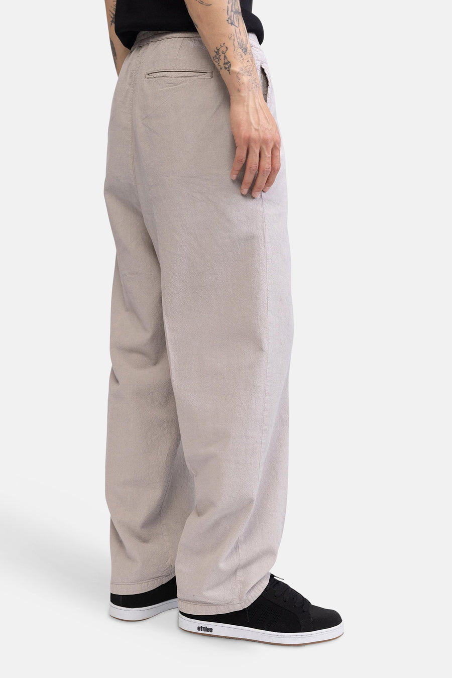 Zijaanzicht van een ecru broek van REDEFINED REBEL, losvallend, met subtiele textuur en achterzak, gedragen met zwarte sneakers.