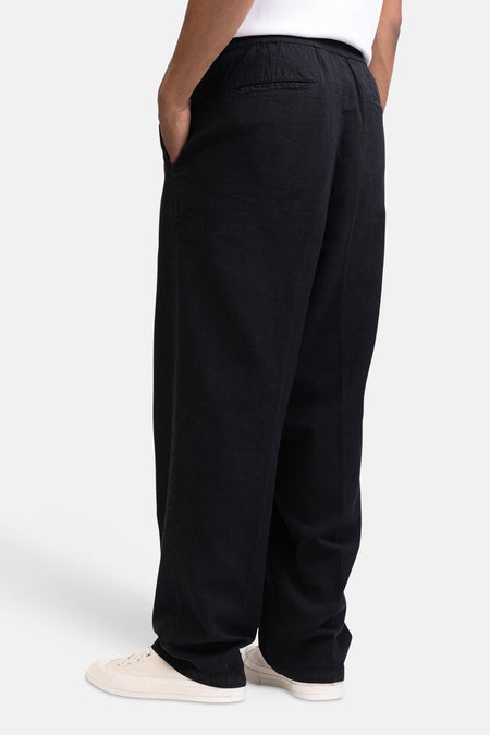 Zwarte broek van REDEFINED REBEL met losse pasvorm, elastische tailleband en hand in zijzak, gedragen met witte t-shirt en beige sneakers.