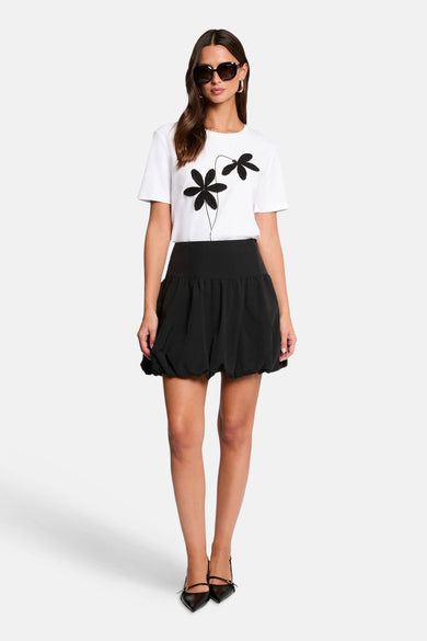 T-shirt blanc à manches courtes de Morgan de Toi, avec imprimé fleurs noires, combiné avec une jupe ballon noire et des chaussures noires.
