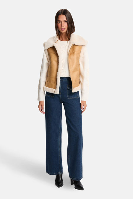 Model in een wit gebreide trui, een bruin suède gilet met schapenvacht en een wijde, donkerblauwe jeans.