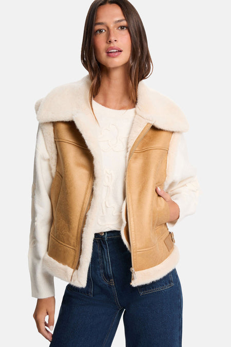 Een vrouw draagt een suède jas met bontvoering, een witte trui en een donkerblauwe jeans tegen een witte achtergrond.