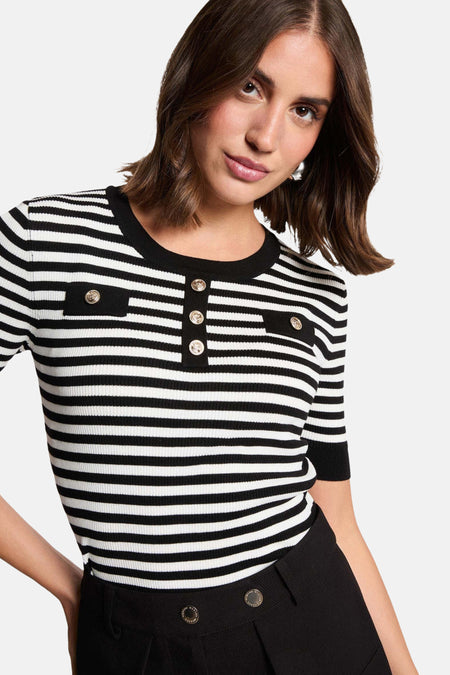 Vrouw in gestreepte top met gouden knopen en zwarte broek.