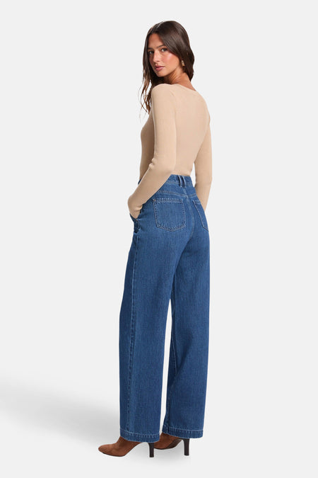 Vrouw draagt wijde jeans en een beige trui, bruine laarsjes, staat in een studio.