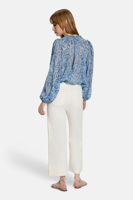 Chemisier bleu à manches longues de Morgan De Toi, avec motif cachemire et encolure froncée, combiné avec un pantalon blanc et des sandales argentées.