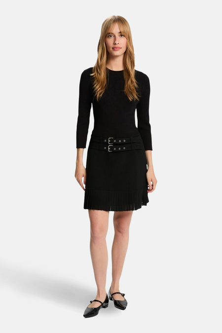 Vrouw in zwarte jurk met plooirok en zwarte schoenen, staand voor witte achtergrond.
